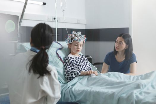 Clinic pediatric medic analyzing EEG scan results of sick little child while in pediatric ward recovery room. Child patient wearing EEG helmet while in pediatric hospital.