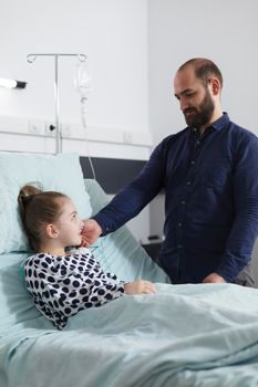 Careful supportive father caress sick little daughter while laying in hospital patient bed. Thoughtful lovely parent offering ill child patient emotional support while resting in patient room.
