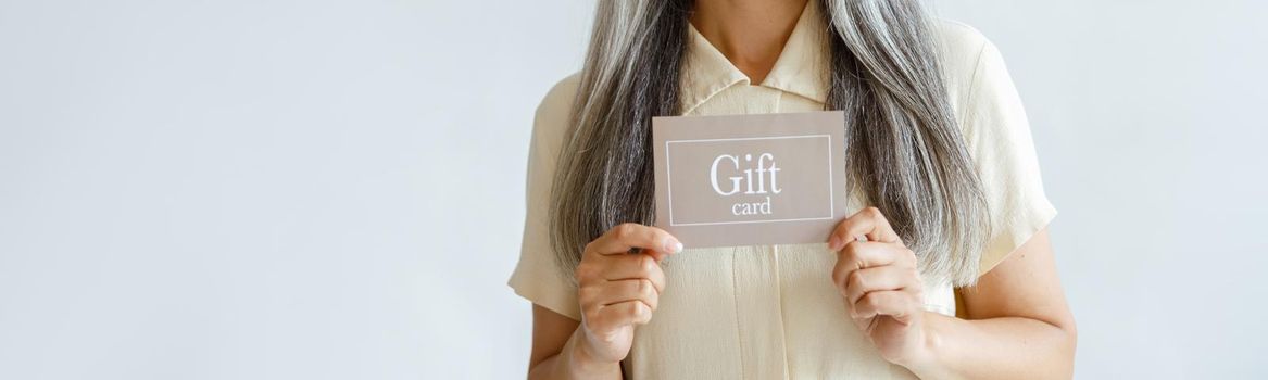 Smiling grey haired Asian woman holds gift card standing on light background