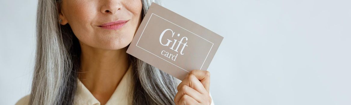 Dreamful grey haired Asian woman holds gift card near face in studio