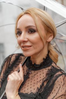 The blonde stands under a transparent umbrella during the rain. The fall season. Rear view. The woman is dressed in a black lace dress, her hair pulled back in a ponytail.