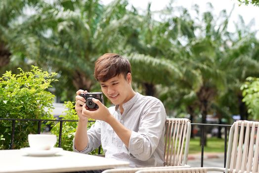 Young serious male sitting at wooden table with photo camera and preparing for photo session while having coffee break