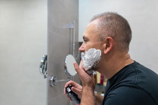 Mature handsome man applying shaving foam on his face to cut off his beard and mustache with razor