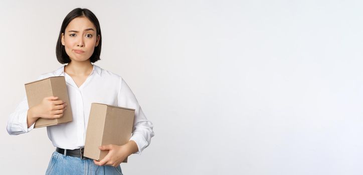 Complicated young asian woman holding two boxes, looking doubtful at camera, standing over white background puzzled