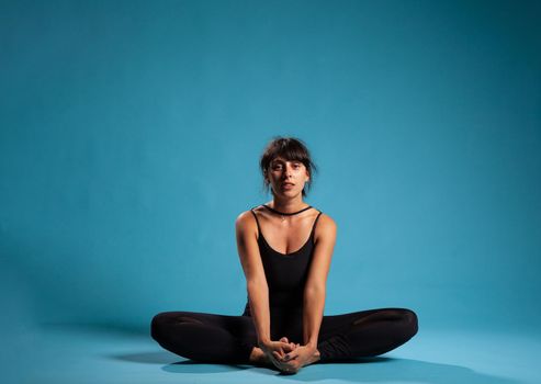 Athletic trainer sitting in padmasana position on yoga mat stretching body muscles in studio with blue background. Athlete woman wearing sportswear practicing lotus posture for healthy lifestyle