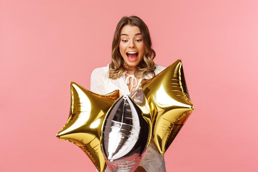 Holidays, celebration and women concept. Portrait of happy lovely young woman in white dress, gasping from amazement and joy, holding birthday star-shaped balloons, pink background