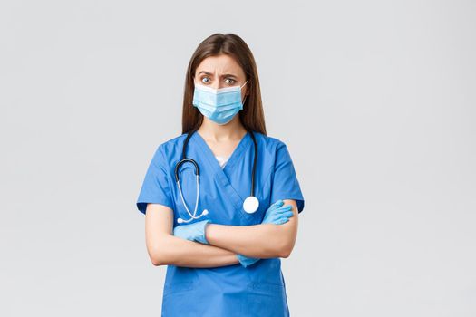 Covid-19, preventing virus, health, healthcare workers and quarantine concept. Skeptical and concerned female nurse in blue scrubs, stethoscope and personal protective equipment look suspicious