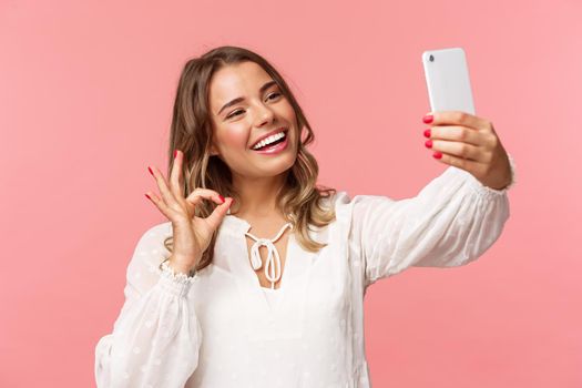 Close-up of satisfied good-looking blond girl in white dress, taking selfie, record mobile phone video, show okay satisfactory sign with pleased nod, smiling agree or recommend, pink background