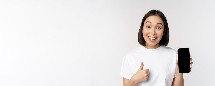 Enthusiastic young woman showing thumb up and mobile phone screen, standing in tshirt over white background