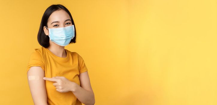 Vaccination and covid-19 pandemic concept. Smiling asian woman in medical face mask, showing her shoulder with band aid after vaccinating from coronavirus, yellow background