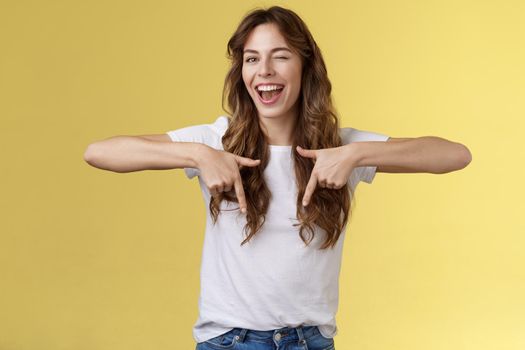 Hey visit my link would not regret. Lively cheeky enthusiastic woman curly long haircut wink sassy smiling broadly show you perfect copy space pointing down bottom promo yellow background