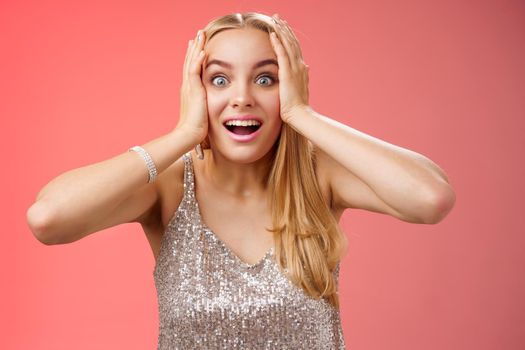 Amused surprised excited young blond attractive woman in silver glittering stylish dress hold head hands widen eyes speechless look camera unbelievable thrill fan see famous person, red background