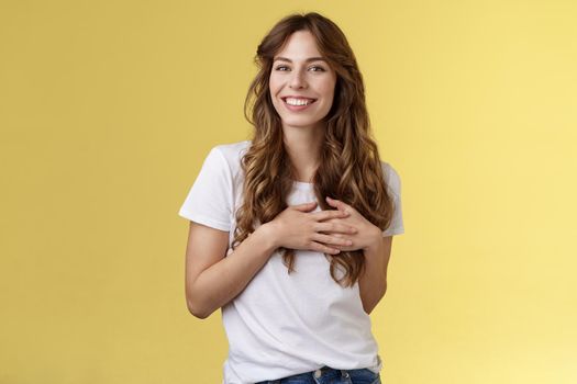 Lovely joyful tender feminine girlfriend long curly hairstyle touch heart feel heartbeat delighted receive lovely heartwarming gift smiling broadly satisfied flirty gazing camera yellow background