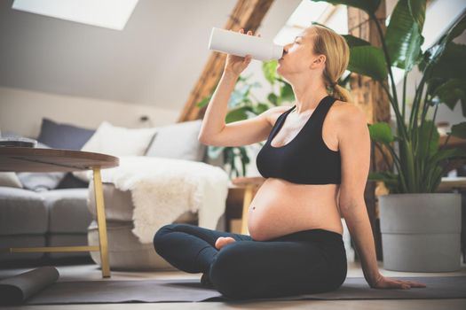 Young happy and cheerful beautiful pregnant woman taking a break, hydrating, drinking water from the botle after home well being workout progrem.