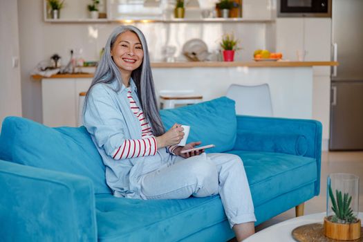 Smiling woman with smartphone and coffee sitting on sofa at home