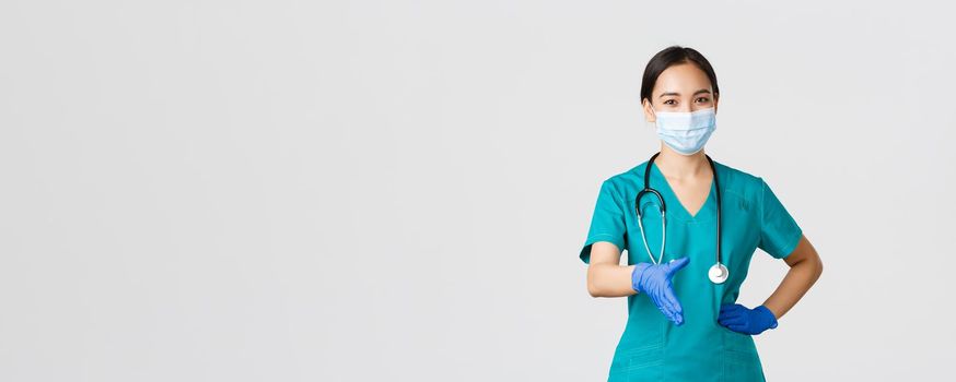 Covid-19, coronavirus disease, healthcare workers concept. Confident young asian female doctor, physician in scrubs and medical mask, gloves, extend hand for handshake, greeting patient