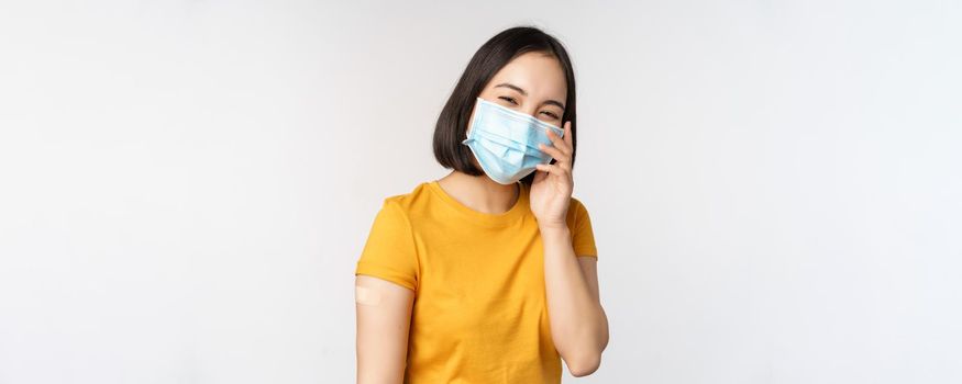 Covid-19, vaccination and healthcare concept. Cute asian girl in medical face mask, showing band aid after coronavirus vaccination, standing over white background