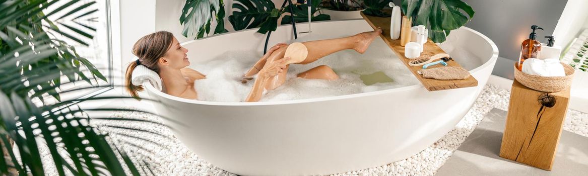 Relaxed young woman using brush while taking buble bath in modern bathroom decorated with tropical plants