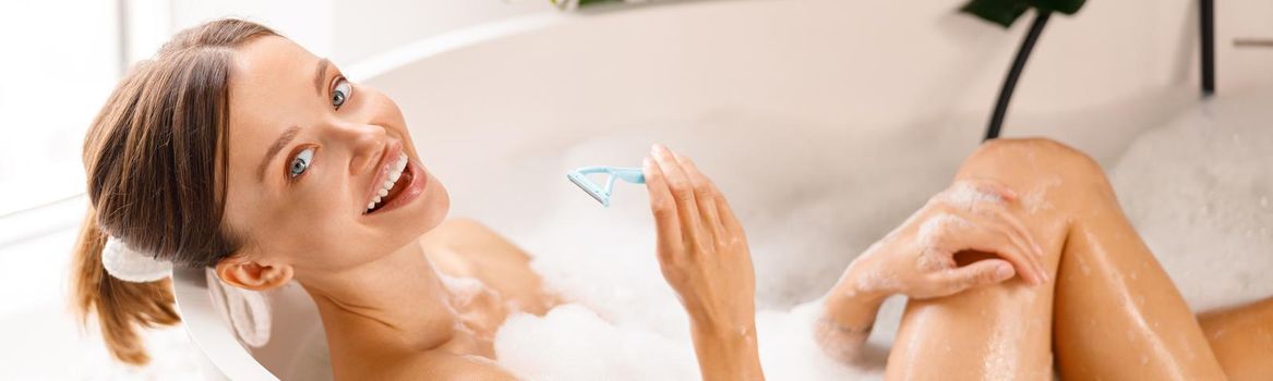 Happy young woman smiling at camera, holding disposable shaving razor for shaving her legs in the bath with foam
