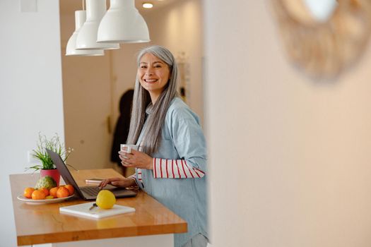 Cheerful woman with cup of coffee using laptop in kitchen