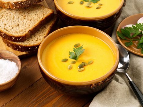 Two brown bowl of pumpkin soup with parsley and pumpkin seeds on brown dark wooden background, side view.