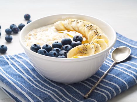 Oatmeal with bananas, blueberries, chia, jam, honey, blue napkin on white wooden background, side view