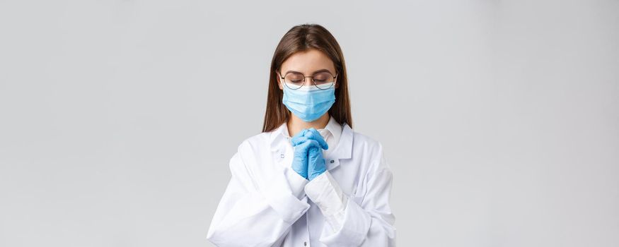 Covid-19, preventing virus, health, healthcare workers and quarantine concept. Hopeful doctor in scrubs and medical mask, personal protective equipment, praying with hands clasped together, hope