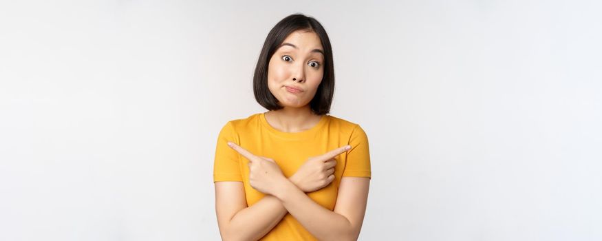 Complicated asian girl pointing fingers sideways, showing left and right choices, staring indecisive clueless, standing over white background