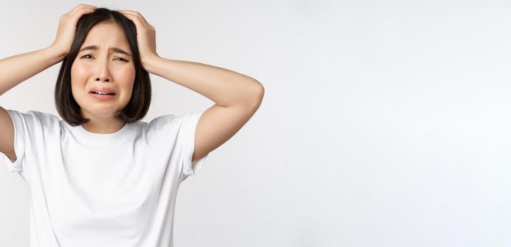 Desperate young korean woman holding hands on head, panicking, crying and standing distressed against white background