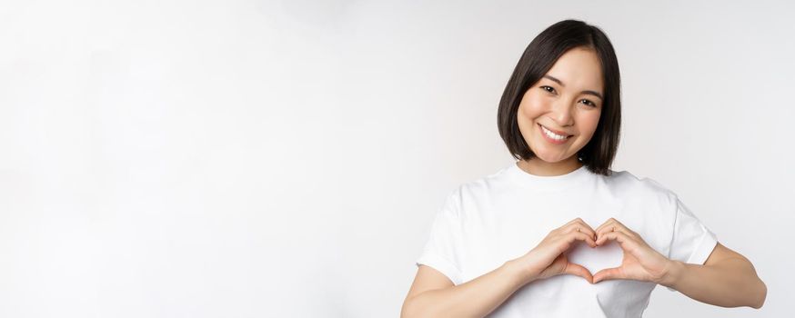 Lovely asian woman smiling, showing heart sign, express tenderness and affection, standing over white background