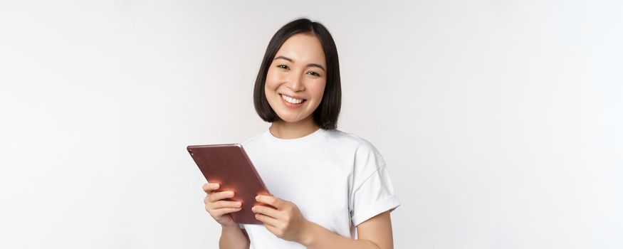 Smiling asian girl with digital tablet, looking happy and laughing, posing in tshirt over white background