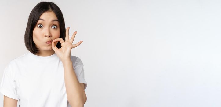 Image of young asian woman showing silence gesture, zipping mouth, seal lips, make promise, standing over white background