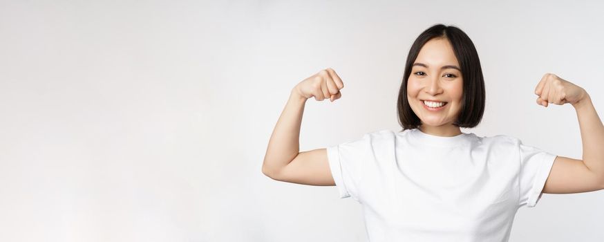 Smiling asian woman showing flexing biceps, muscles strong arms gesture, standing in white tshirt over white background