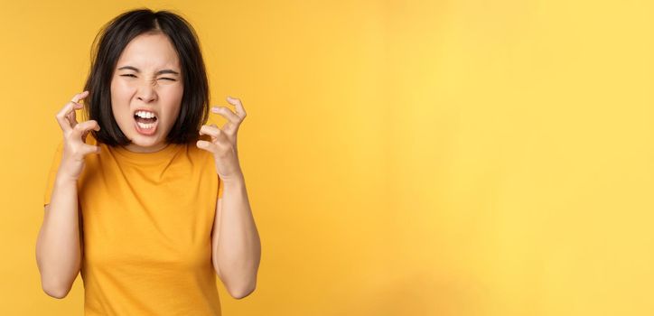 Image of angry asian woman, shouting and cursing, looking outraged, furious face expression, standing over yellow background