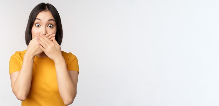 Shocked asian woman cover mouth with hands, looking startled with speechless face expression, standing in yellow tshirt against white background