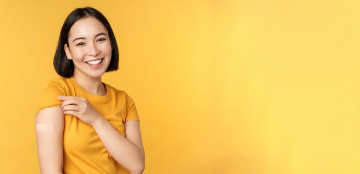 Vaccination and covid-19 pandemic concept. Happy and healthy asian girl pointing at her shoulder with band aid after vaccinating from coronavirus, yellow background