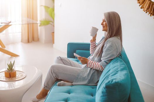 Smiling woman drinking coffee and using cellphone at home