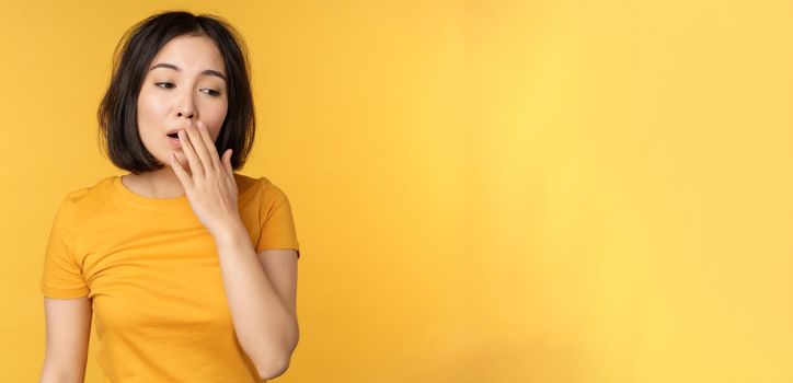 Image of young asian girl student, yawning, feeling tired, standing sleepy agaisnt yellow background