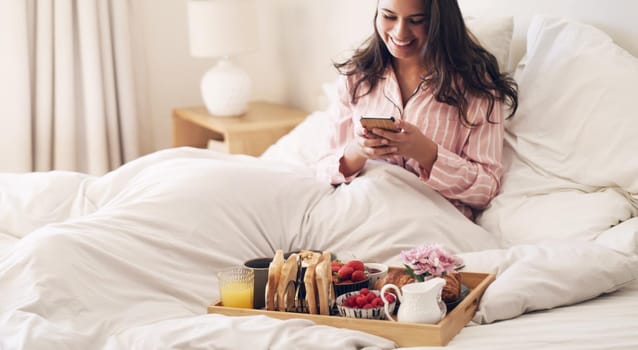 The best way to start the day. Shot of a beautiful young woman using a smartphone while having breakfast in bed at home.