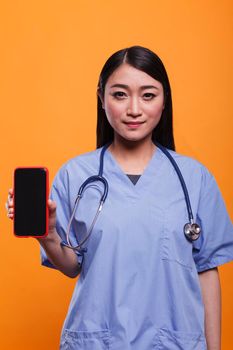 Attractive healthcare clinic nurse wearing stethoscope while showing smartphone device to camera.. Hospital caregiver wearing medical instrument while holding mobile cellphone on orange background.