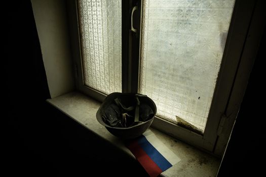Conceptual photo of war between Russia and Ukraine. Russian helmet and flag on windowsill at night. Old creepy room with window. Explosion outside.