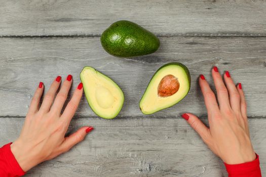 Tabletop view - woman hands with red nails, avocado cut in half, whole one above, on gray wood desk.