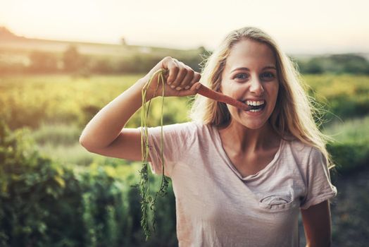 Delicious and nutritious. Cropped portrait of a young woman holding up a carrot and taking a bite with her farmland in the background.