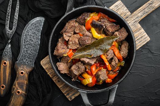 Beef meat stewed with potatoes, carrots and spices, in cast iron frying pan, on black stone background, top view flat lay
