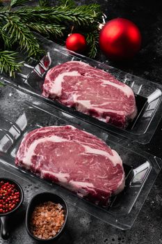 Rib-eye beef steak in plastic packing tray with Christmas tree decorations, on black dark stone table background