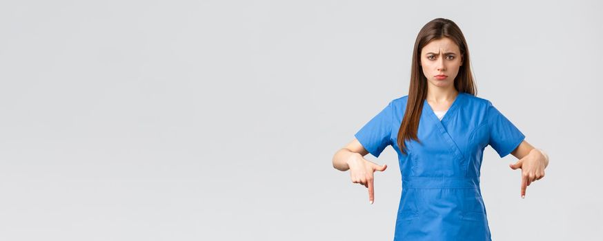 Healthcare workers, prevent virus, covid-19 test screening, medicine concept. Disappointed and uneasy cute nurse or doctor in blue scrubs, pointing fingers down, showing bad unpleasant news