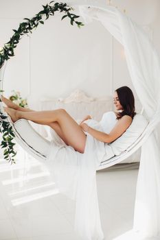 Gorgeous pregnant woman in white gown on designed chair.