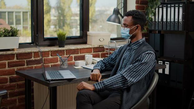 Employee with face mask using video call communication on laptop to talk to colleagues about business project. Man talking to people on online teleconference, having remote conversation.