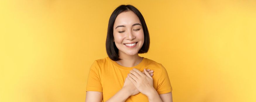 Beautiful asian woman, smiling with tenderness and care, holding hands on heart, standing in tshirt over yellow background