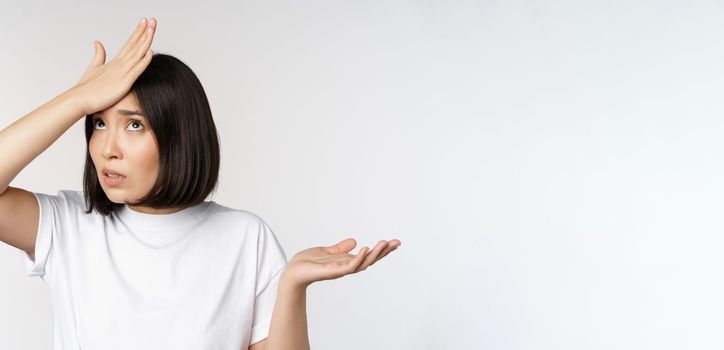Annoyed korean girl facepalm, slap forehead and shrugging, confused by smth, standing over white background